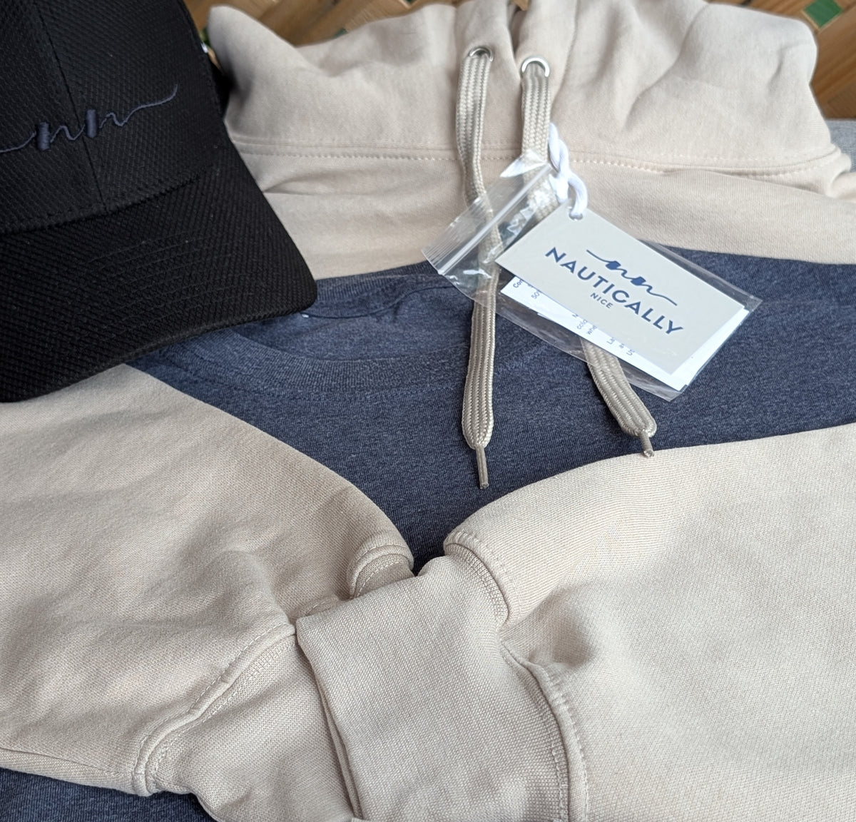 Nautically Nice brand hang tag resting on a Dunedrift hoodie and Moormist tee alongside the embroidered cap